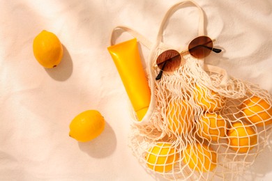Photo of String bag with sunglasses, lemons and sunscreen on sand, flat lay