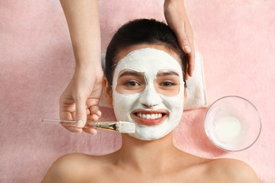 Cosmetologist applying white mask onto woman's face in spa salon, top view