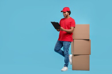 Photo of Happy young courier with clipboard and stack of parcels on light blue background, space for text