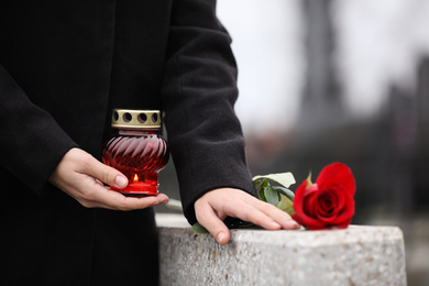 Woman holding candle near grey granite tombstone outdoors, closeup. Funeral ceremony