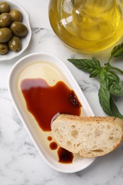 Bowl of organic balsamic vinegar with oil served with bread slice, basil and olives on white marble table, flat lay