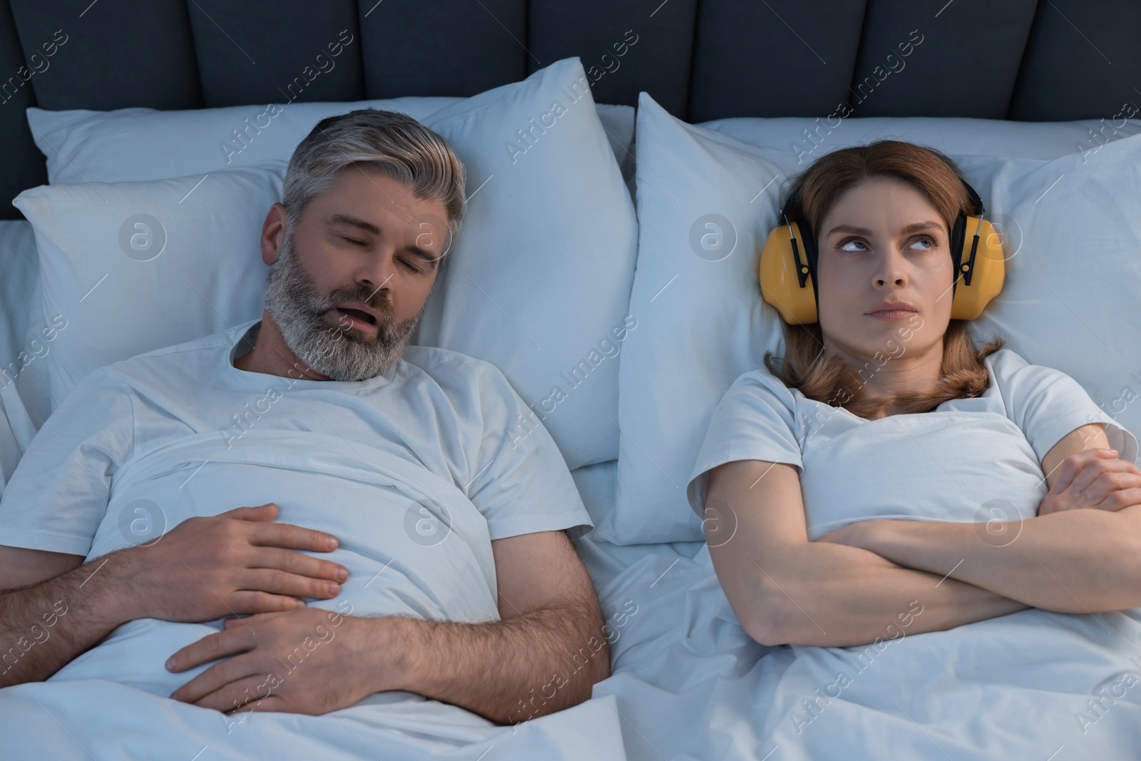 Photo of Irritated woman with headphones lying near her snoring husband in bed at home