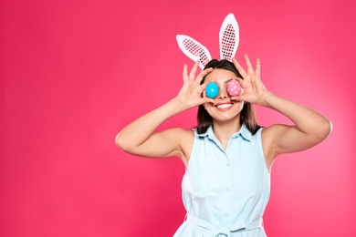 Photo of Beautiful woman in bunny ears headband holding Easter eggs near eyes on color background, space for text