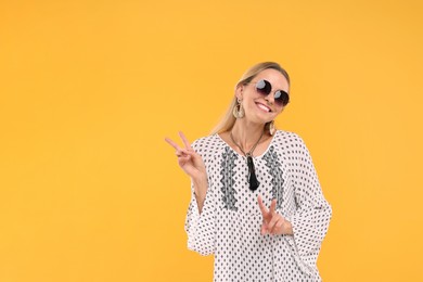 Portrait of smiling hippie woman showing peace signs on yellow background. Space for text