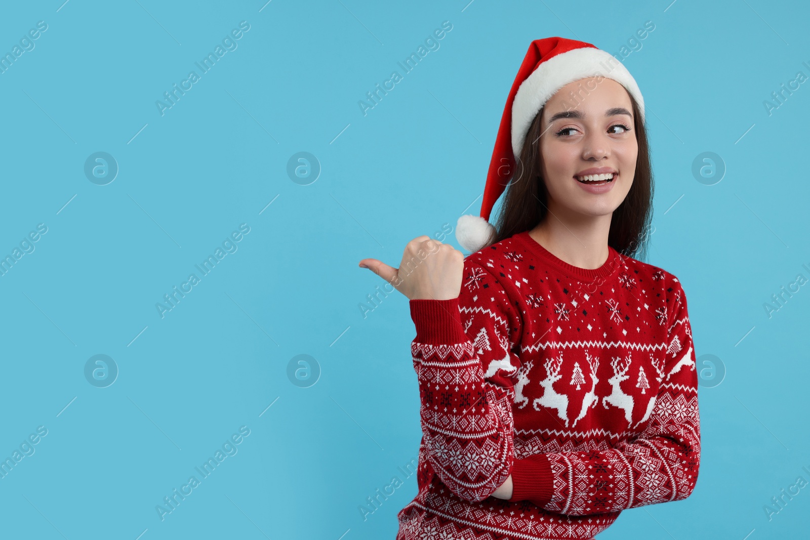 Photo of Happy young woman in Christmas sweater and Santa hat pointing at something on light blue background. Space for text