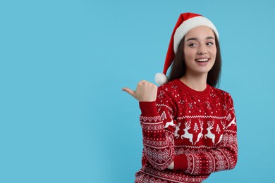 Happy young woman in Christmas sweater and Santa hat pointing at something on light blue background. Space for text