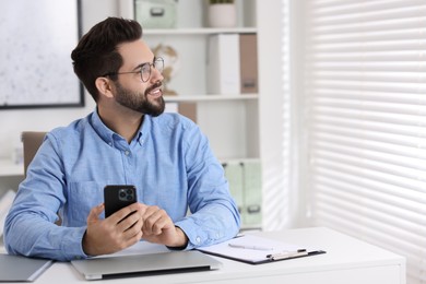 Happy young man using smartphone at white table in office, space for text