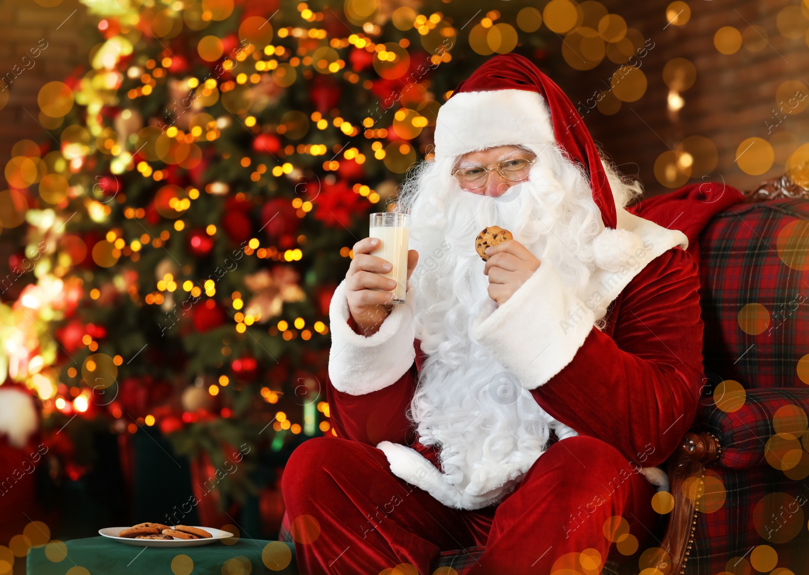 Photo of Santa Claus with milk and cookie near Christmas tree indoors