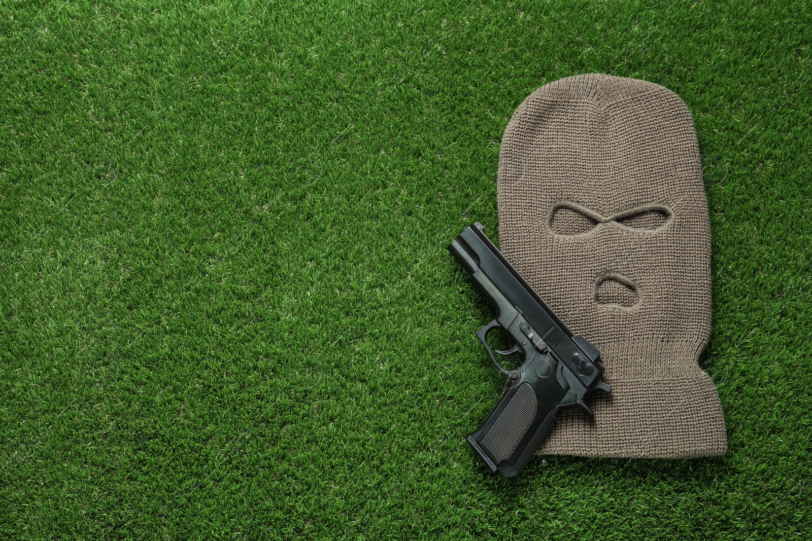 Photo of Beige knitted balaclava and gun on green grass, flat lay. Space for text