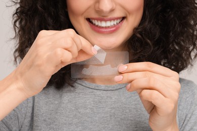 Photo of Young woman holding teeth whitening strips on light grey background, closeup