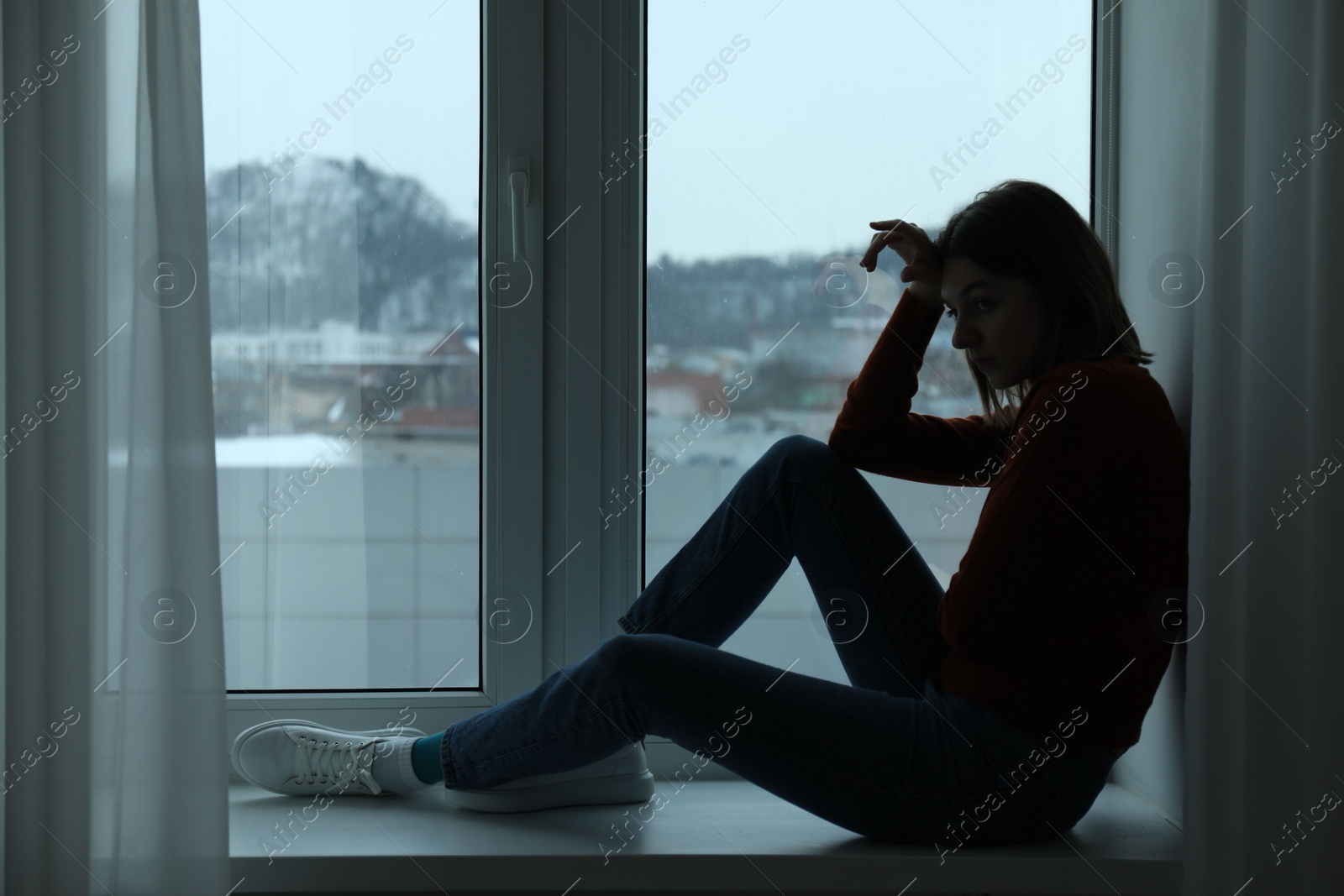 Photo of Sad young woman sitting on windowsill near window at home