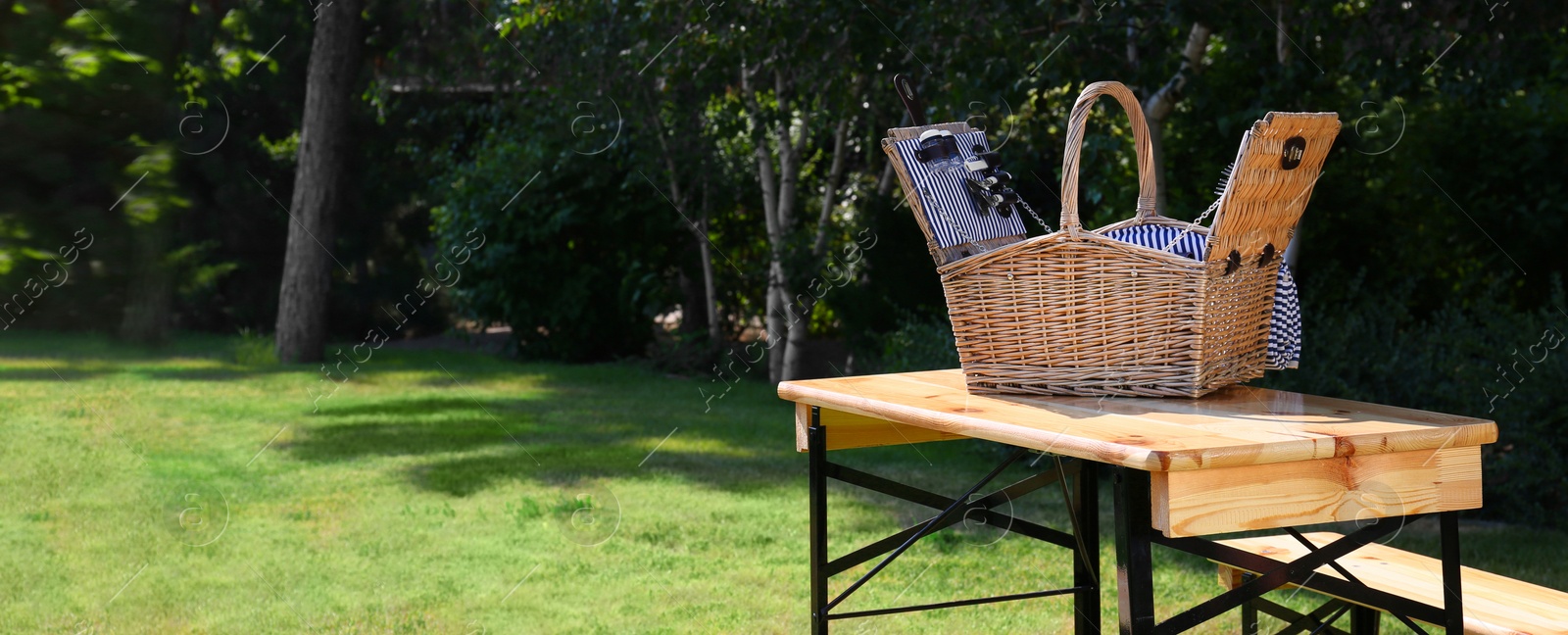 Image of Open picnic basket on wooden table in green park, space for text. Banner design