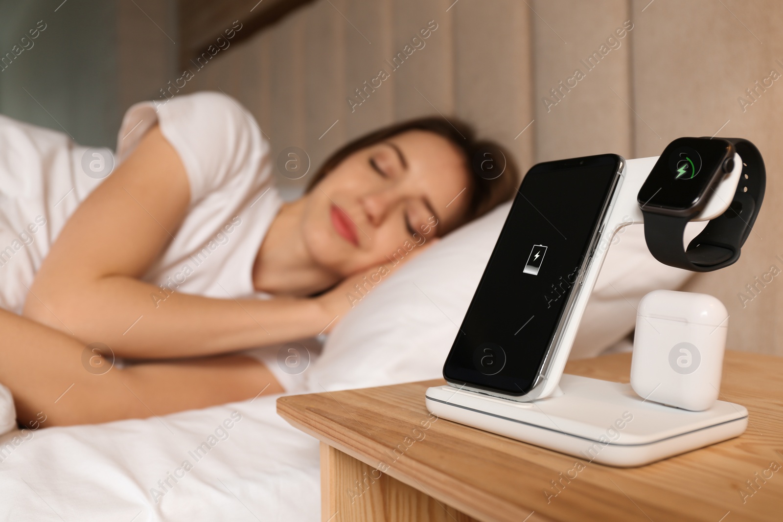 Photo of Smartphone, watch, earphones charging on wireless pad and sleeping woman in room