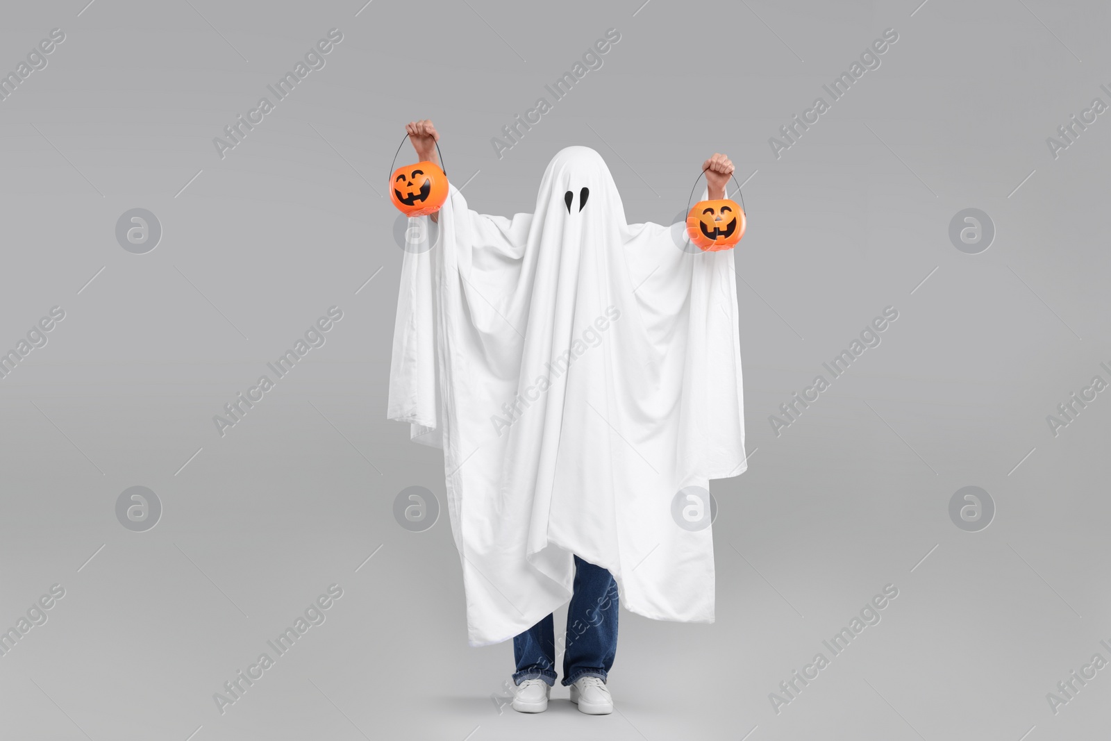 Photo of Woman in white ghost costume holding pumpkin buckets on light grey background. Halloween celebration