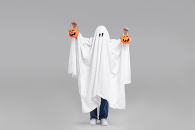 Woman in white ghost costume holding pumpkin buckets on light grey background. Halloween celebration