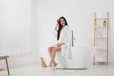 Photo of Beautiful happy woman in stylish bathrobe sitting on tub in bathroom