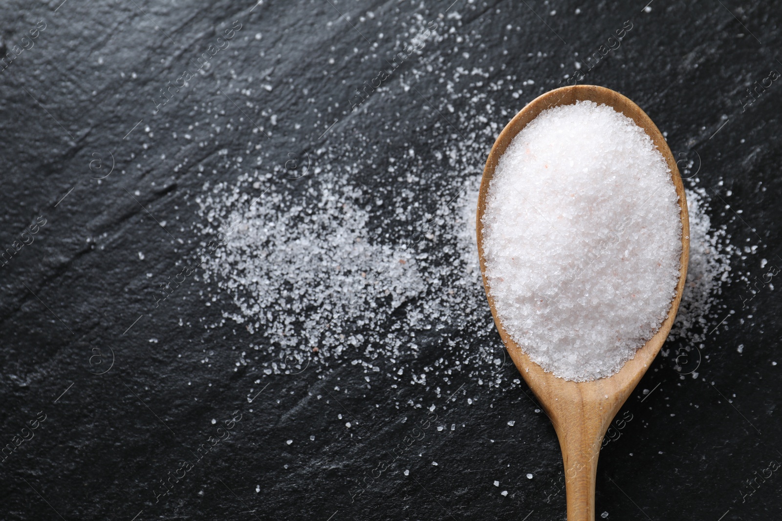 Photo of Organic white salt in spoon on black table, top view. Space for text