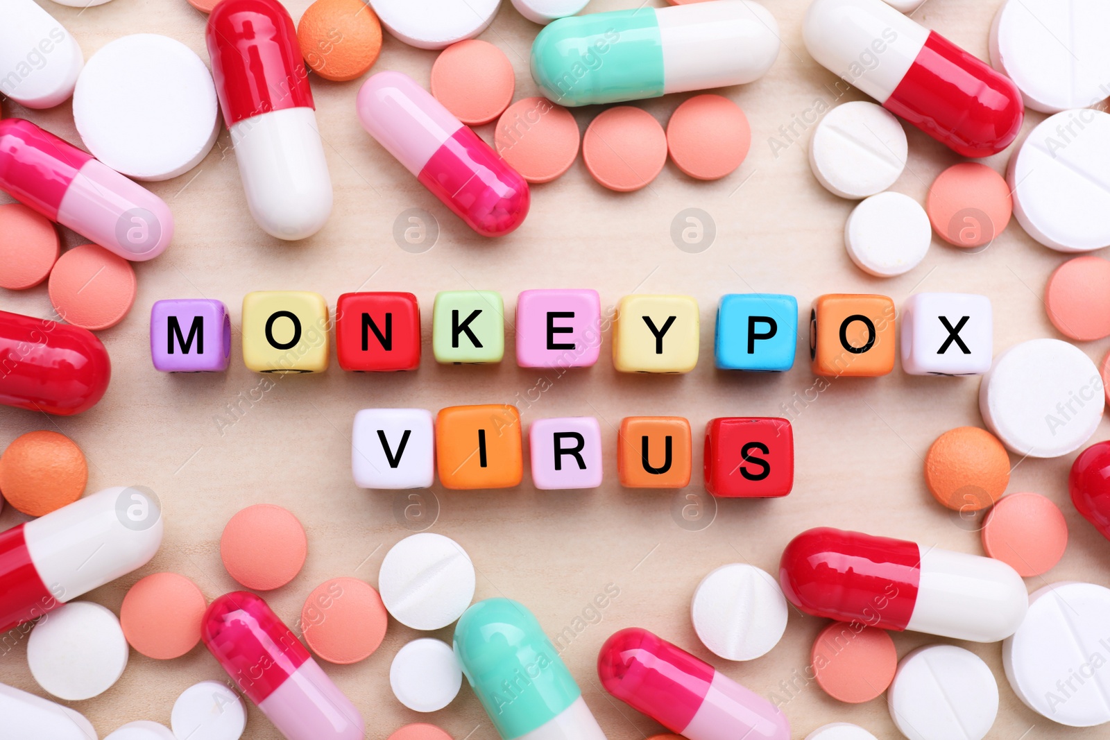 Photo of Words Monkeypox Virus made of colorful cubes surrounded by different pills on wooden table, flat lay