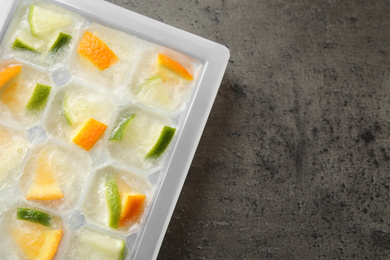 Ice cube tray with frozen fruits on grey table, flat lay. Space for text