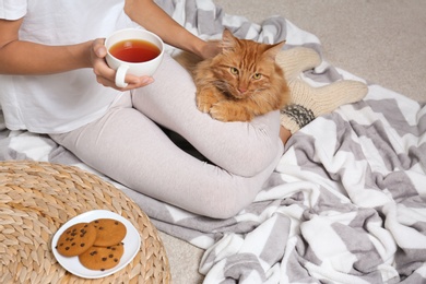 Woman with cute red cat and cup of tea on blanket at home, closeup view