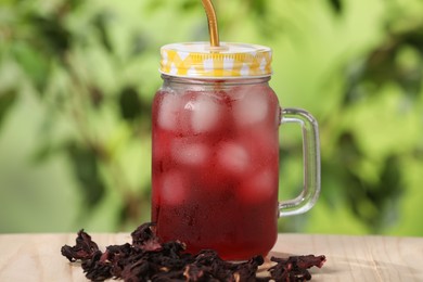 Photo of Refreshing hibiscus tea with ice cubes in mason jar and dry roselle flowers on wooden table against blurred green background