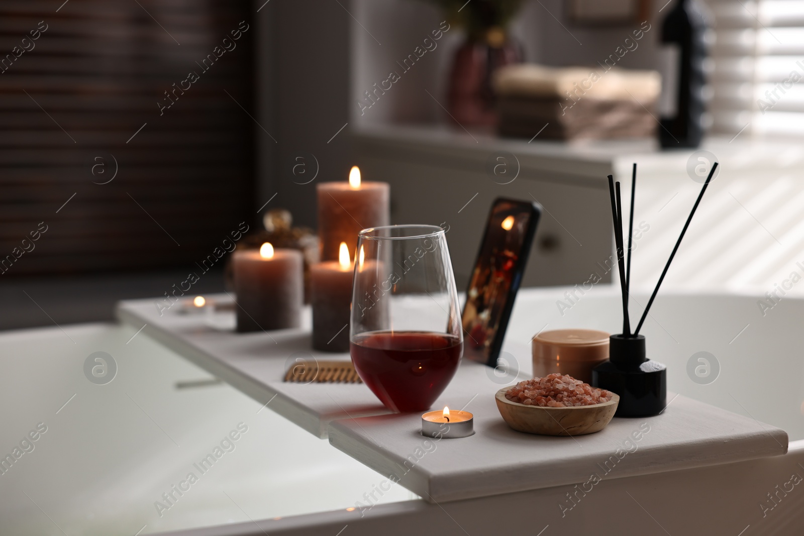 Photo of White wooden tray with glass of wine, beauty products and burning candles on bathtub in bathroom