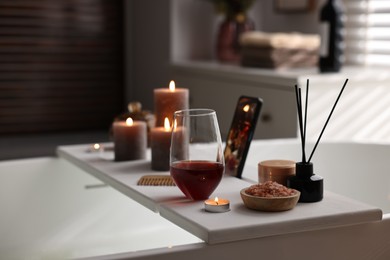 White wooden tray with glass of wine, beauty products and burning candles on bathtub in bathroom