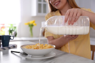 Young woman pouring milk from gallon bottle into plate with breakfast cereal at white marble table in kitchen, closeup