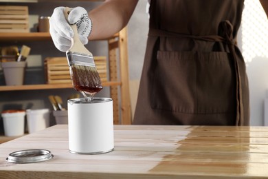 Photo of Man dipping brush into can with varnish at wooden table indoors, closeup