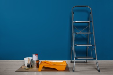 Photo of Metallic folding ladder and painting tools near blue wall indoors, space for text