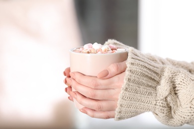 Photo of Woman holding cup of aromatic cacao with marshmallows on blurred background, closeup. Space for text