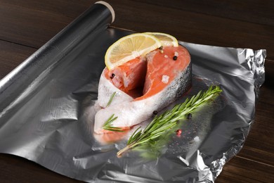 Photo of Aluminum foil with raw fish, lemon slices, rosemary and spices on wooden table. Baking salmon