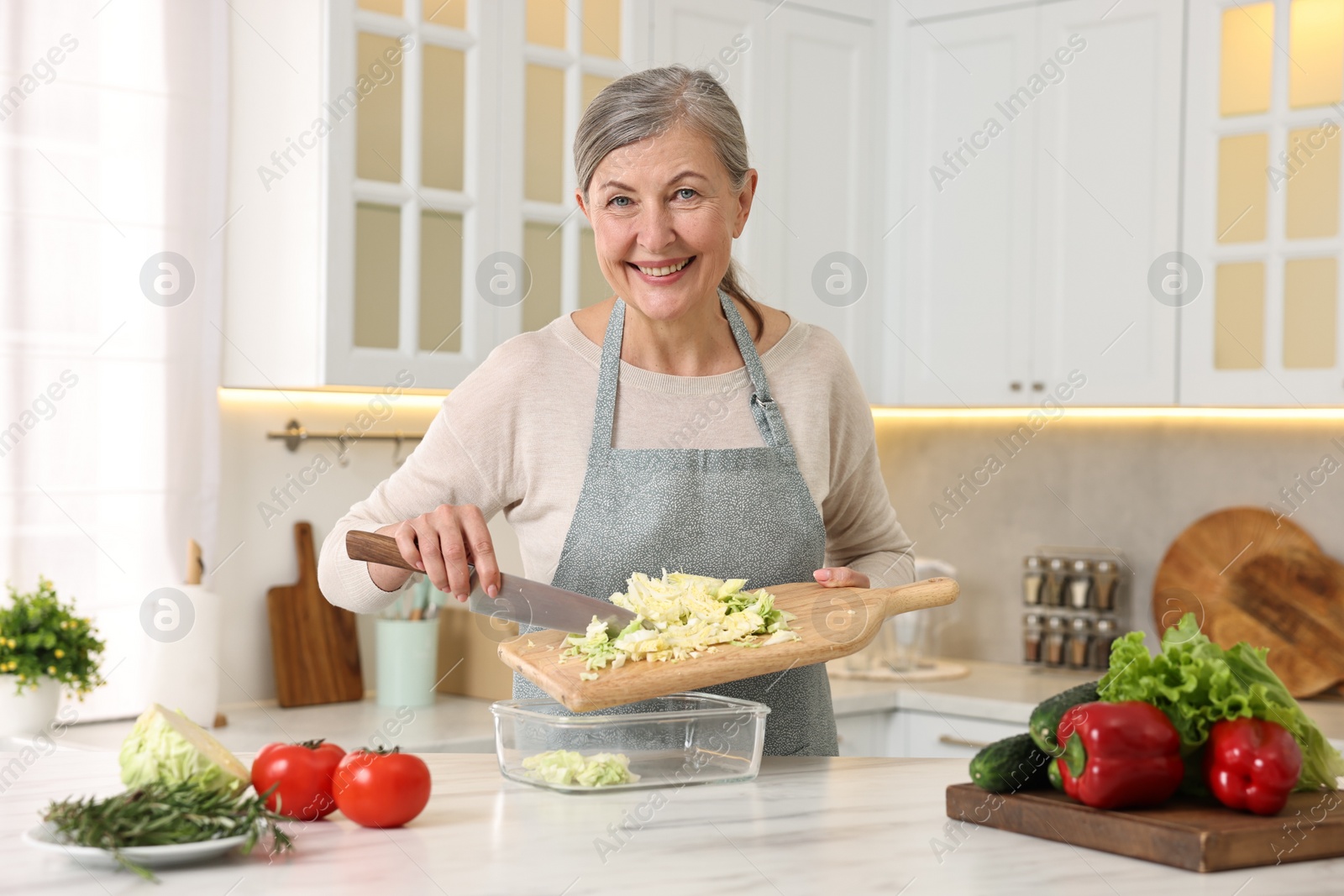 Photo of Happy housewife putting cut cabbage into glass container at white marble table in kitchen