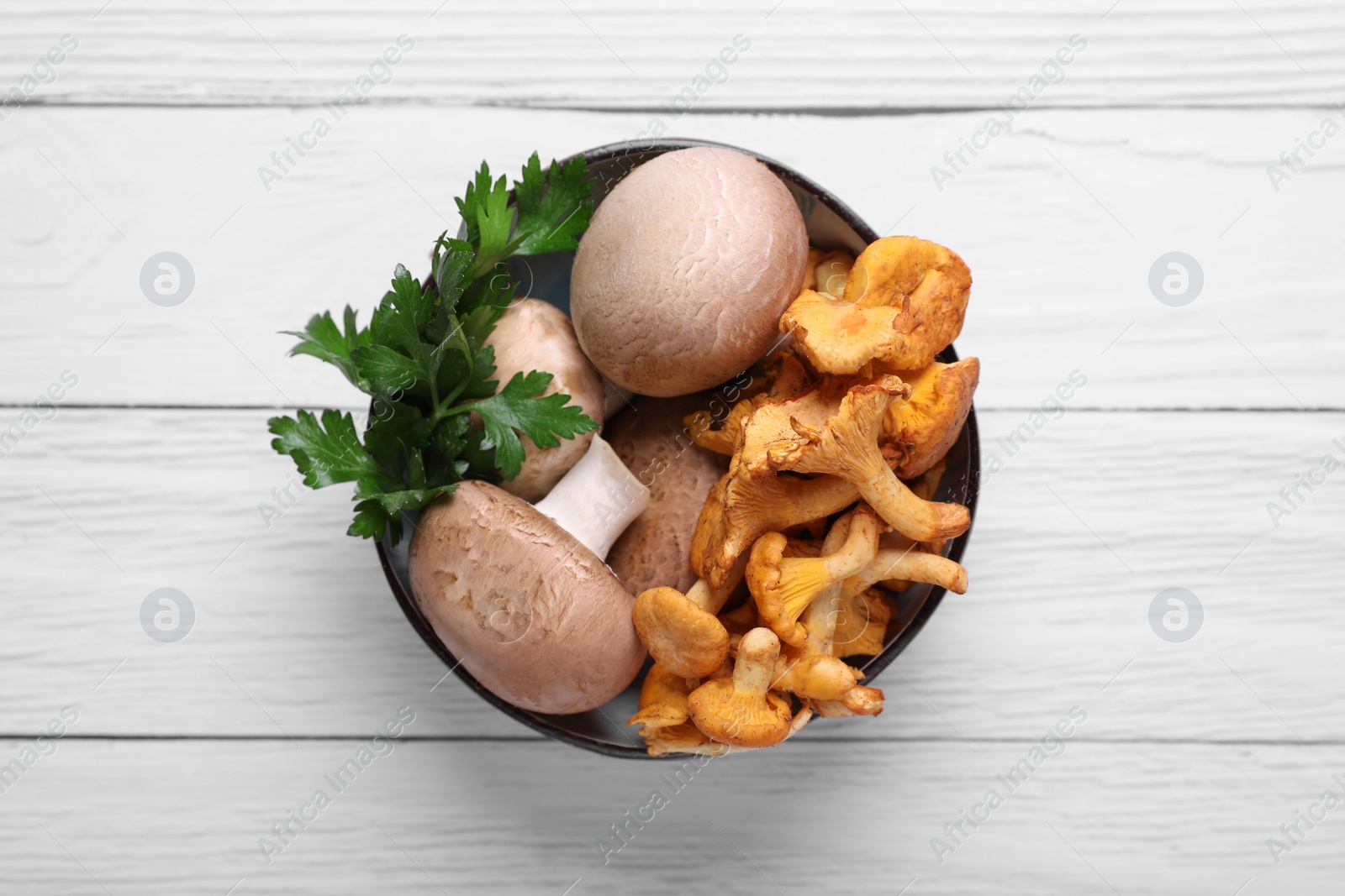 Photo of Bowl of different mushrooms and parsley on white wooden table, top view