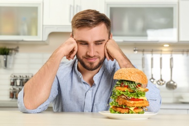 Young man near huge burger on table in kitchen