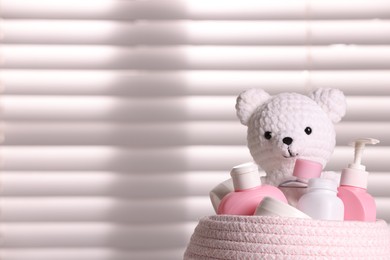 Wicker basket with baby cosmetic products and knitted toy bear near window, closeup. Space for text