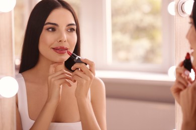Photo of Young woman applying beautiful glossy lipstick in front of mirror indoors