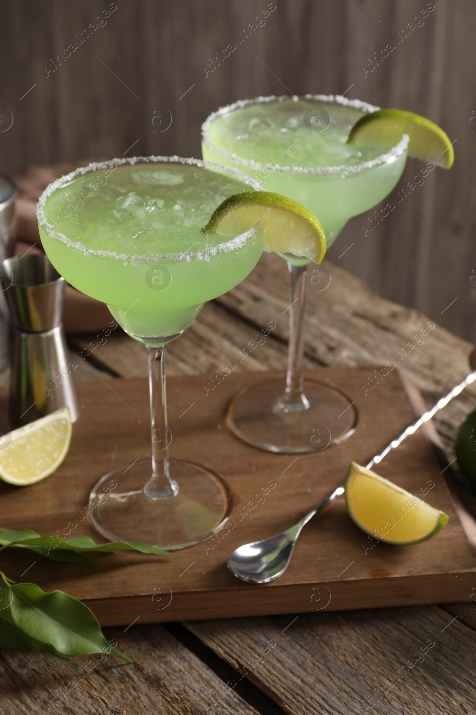 Photo of Delicious Margarita cocktail in glasses, lime, green leaves and bar spoon on wooden table