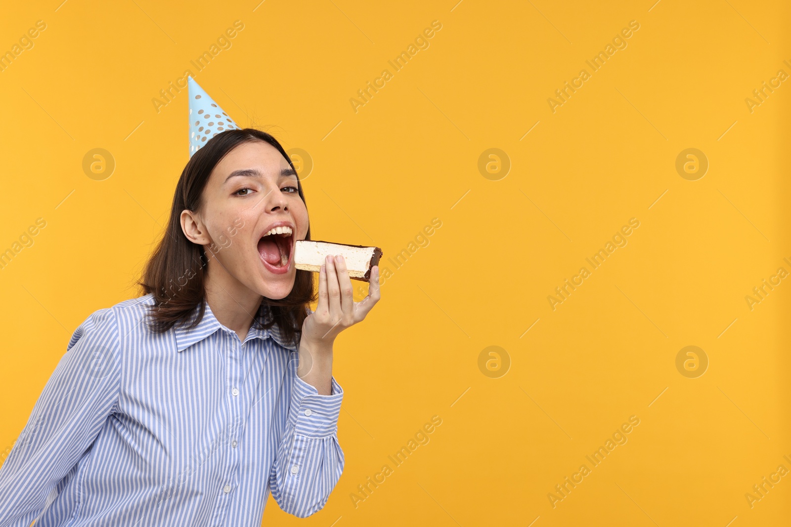 Photo of Happy young woman in party hat eating cheesecake on yellow background