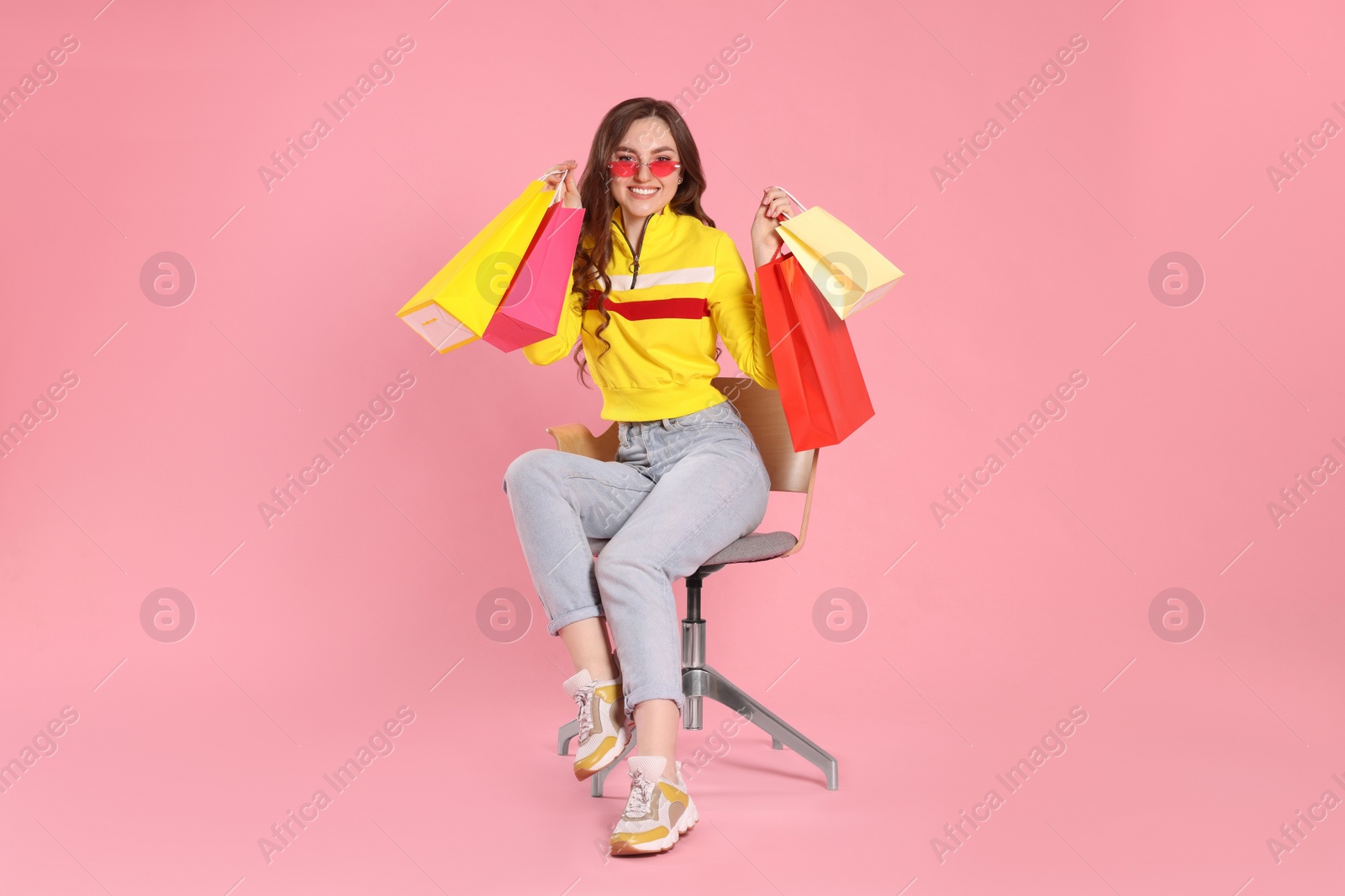 Photo of Happy woman in stylish sunglasses holding many colorful shopping bags on armchair against pink background