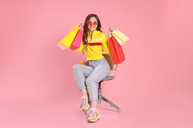 Happy woman in stylish sunglasses holding many colorful shopping bags on armchair against pink background
