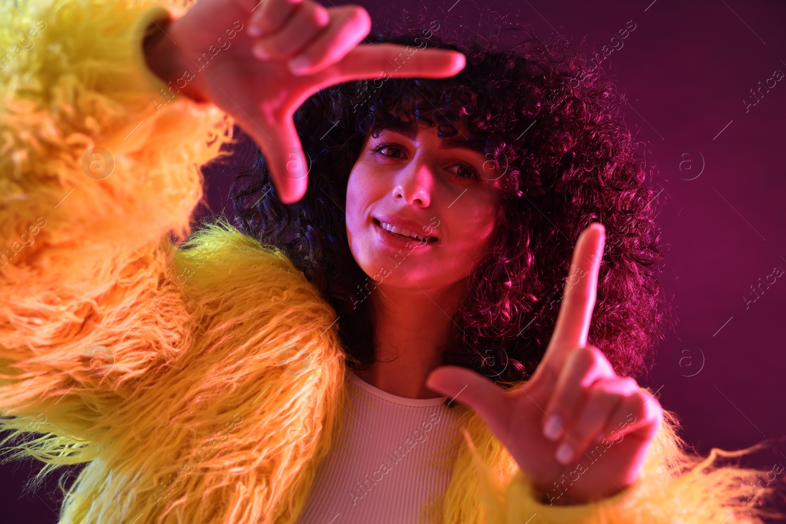 Photo of Beautiful young woman in yellow fur coat making frame gesture with hands on color background in neon lights