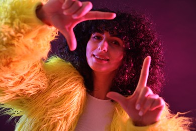 Beautiful young woman in yellow fur coat making frame gesture with hands on color background in neon lights