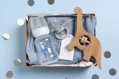 Different baby accessories with blank card in box and confetti on light blue background, top view