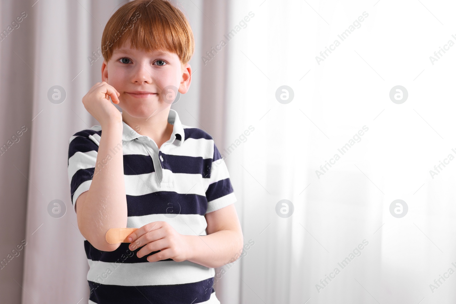 Photo of Little boy putting sticking plaster onto elbow indoors. Space for text