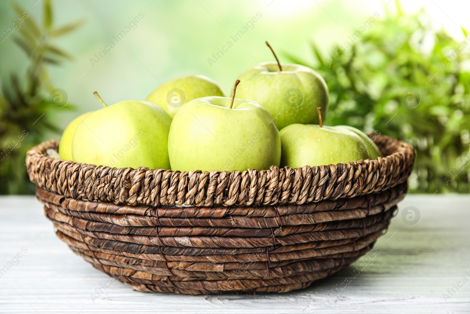 Photo of Wicker bowl of fresh ripe green apples on white wooden table against blurred background