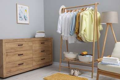 Wardrobe organization. Rack with different stylish clothes, chest of drawers, armchair and lamp near grey wall indoors