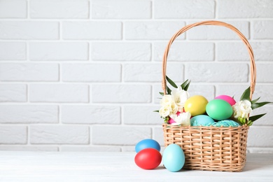 Photo of Wicker basket with bright painted Easter eggs and spring flowers on white wooden table near brick wall. Space for text