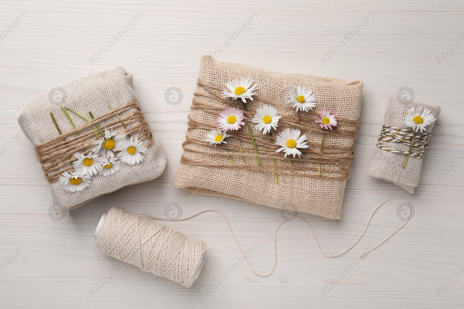 Photo of Gifts packed in burlap fabric with beautiful chamomiles, hemp rope and spool of threads on white wooden table, flat lay