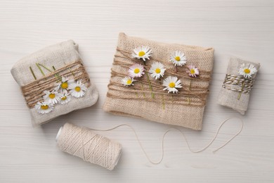 Gifts packed in burlap fabric with beautiful chamomiles, hemp rope and spool of threads on white wooden table, flat lay